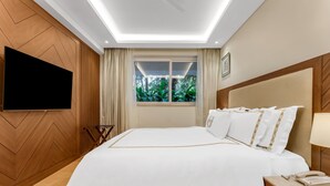 A hotel room with a large bed, a flat-screen TV mounted on the wall, a window with curtains, and a bedside table with a lamp and phone. - Masaka Suites A Unit Of Arbat Hospitality LLP (Betalbatim)