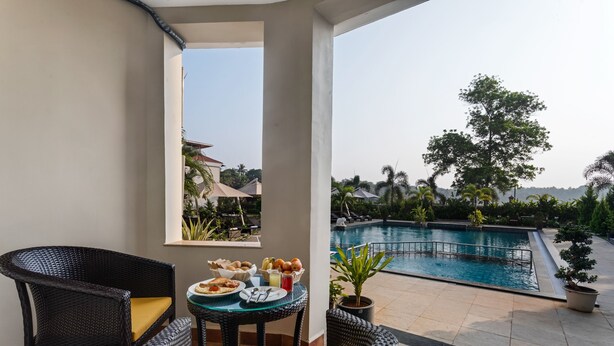 A balcony with wicker furniture, a dining table with food, and a pool view. - Masaka Suites A Unit Of Arbat Hospitality LLP (Betalbatim)