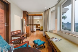 A modern hotel room with a wooden dining area, a cozy seating area, and a large window offering a view of the outdoors. - Masaka Suites A Unit Of Arbat Hospitality LLP (Betalbatim)