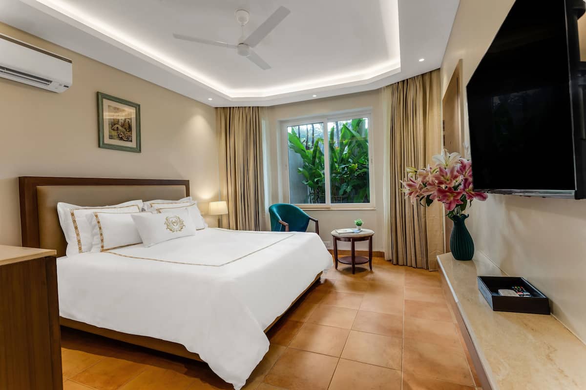 A hotel room with a large bed, a ceiling fan, a TV, a window with curtains, a small table with a vase, and a plant.