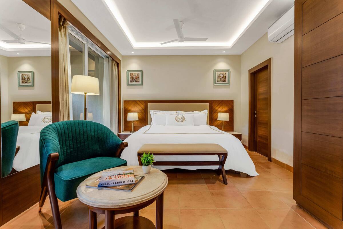 A hotel room with a large bed, a green armchair, a small round table with a magazine, and a potted plant.