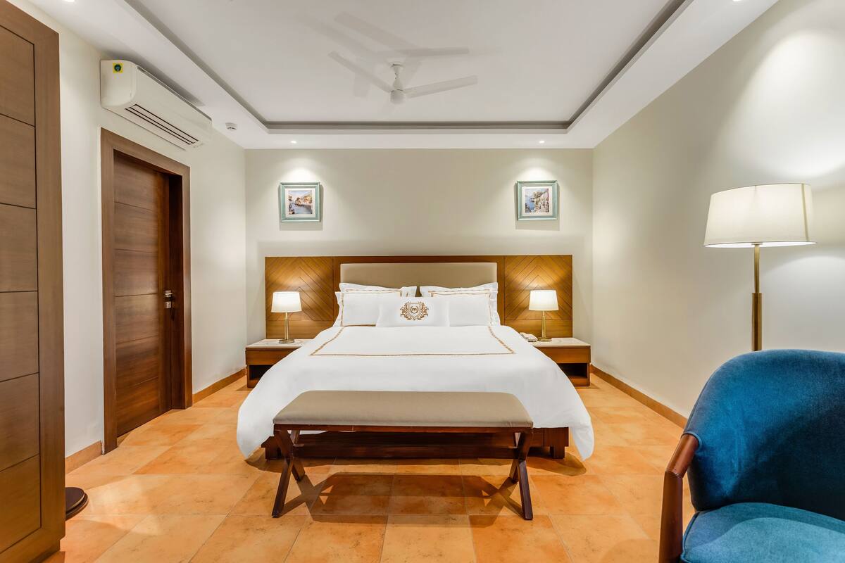 A hotel room with a large bed, two bedside tables with lamps, a bench, a blue chair, and a ceiling fan.
