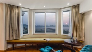 A corner room with a bench, two chairs, a table with a bottle and glasses, and a view through large windows. - Masaka Suites A Unit Of Arbat Hospitality LLP (Betalbatim)