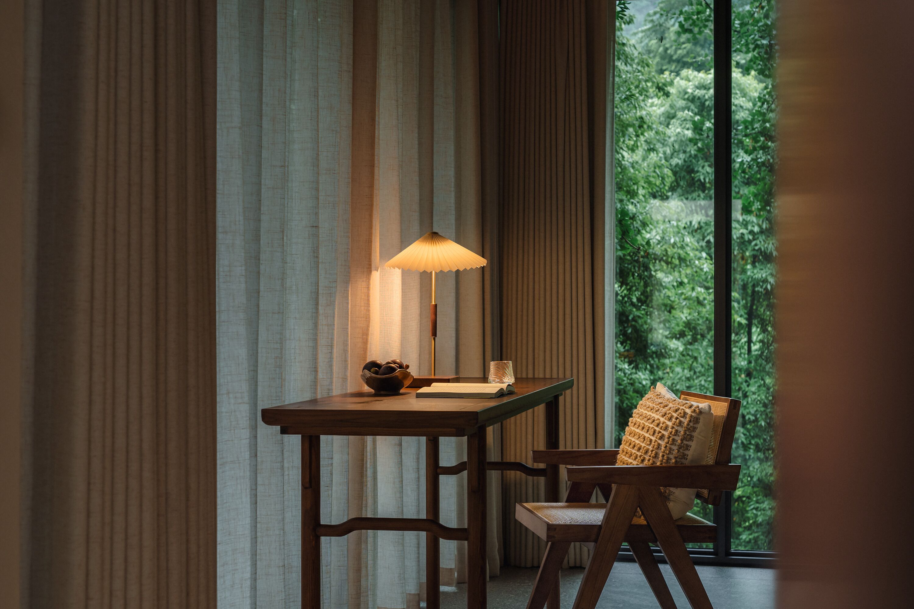 A wooden desk with a lamp, a bowl, and a book, positioned next to a window with curtains.