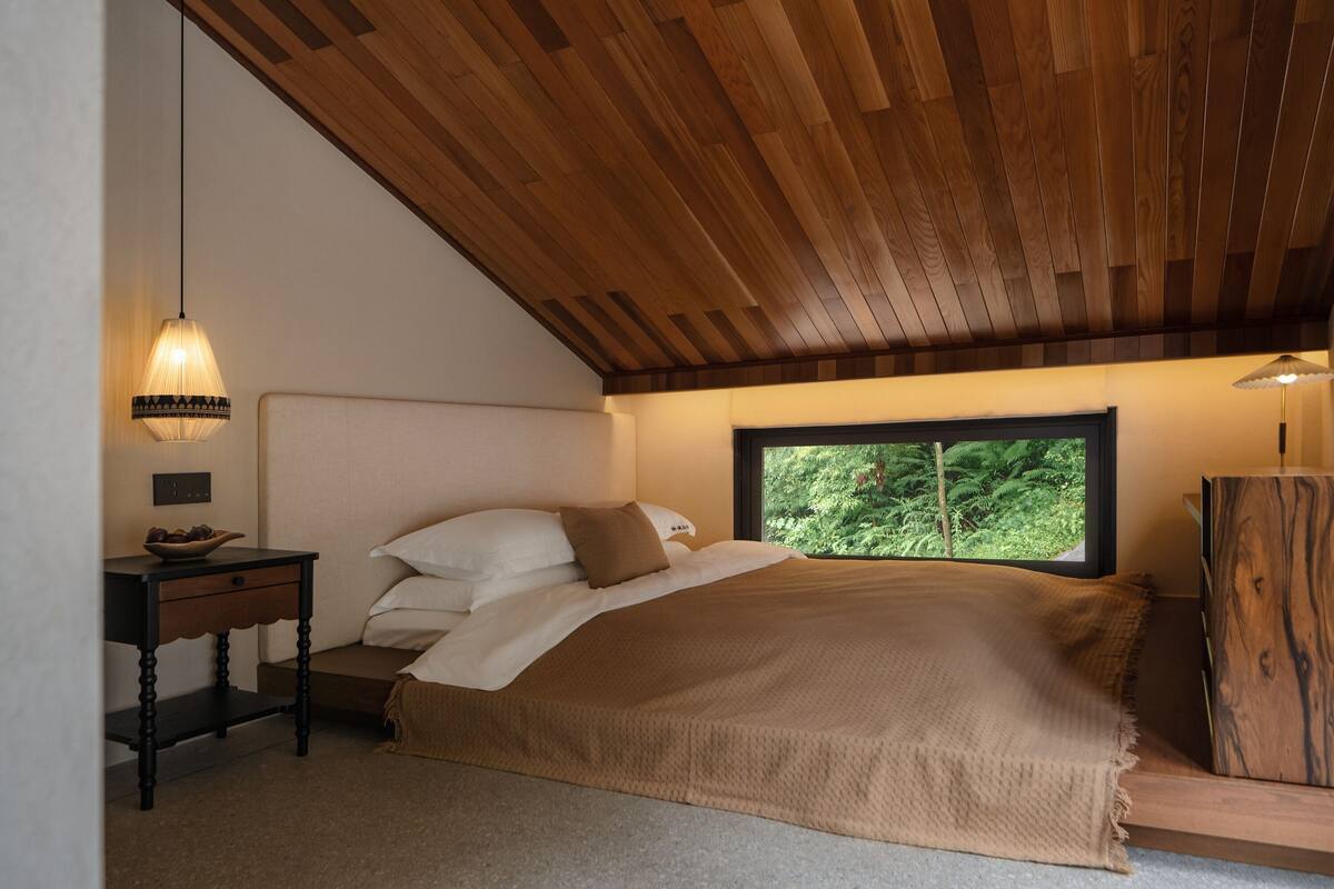 A bedroom with a wooden ceiling, a bed with a beige blanket, a nightstand with a bowl, and a window with a view of greenery.