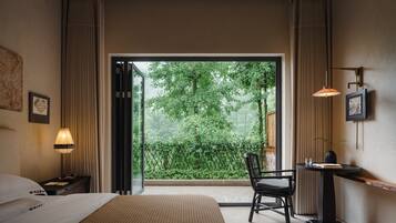 A hotel room with a bed, a desk with a chair, a window with a view of greenery, and a wall-mounted lamp.