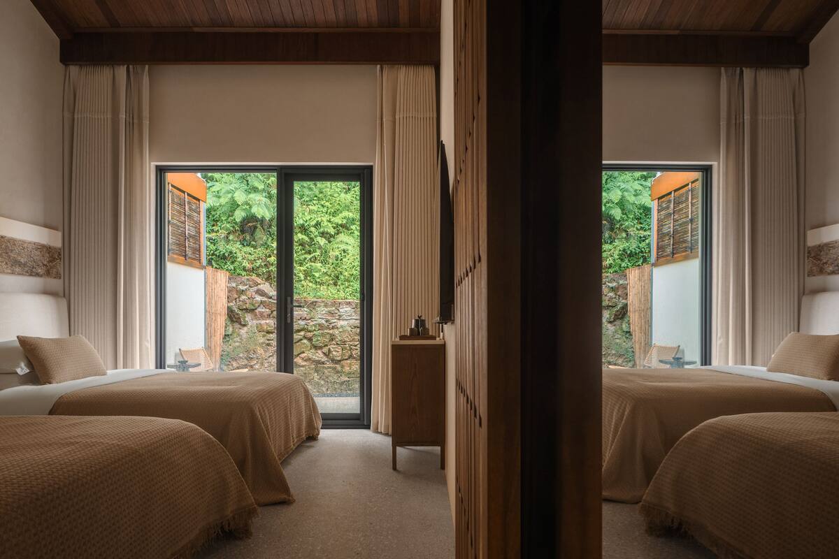 A hotel room with two beds, a wooden ceiling, and a view of a garden through a glass door.