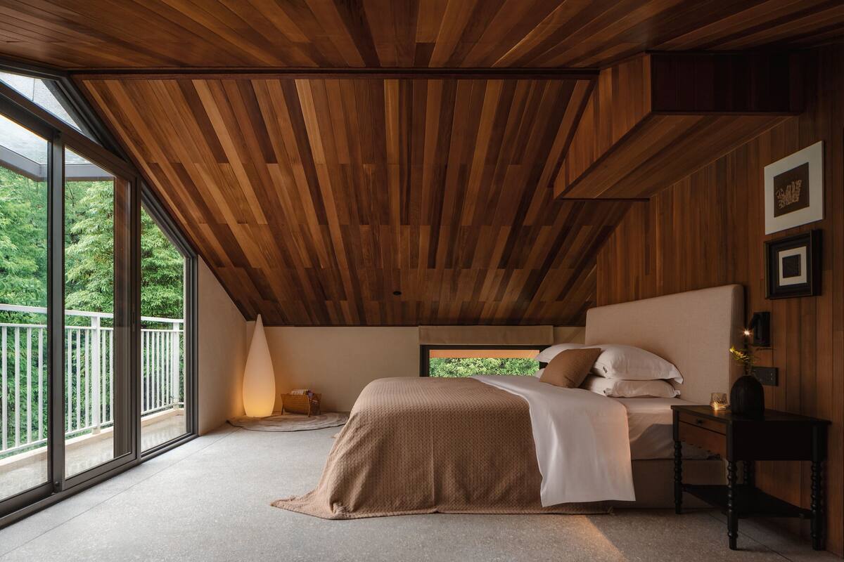 A bedroom with a sloped ceiling, a bed, a bedside table, and a window with a view of greenery.