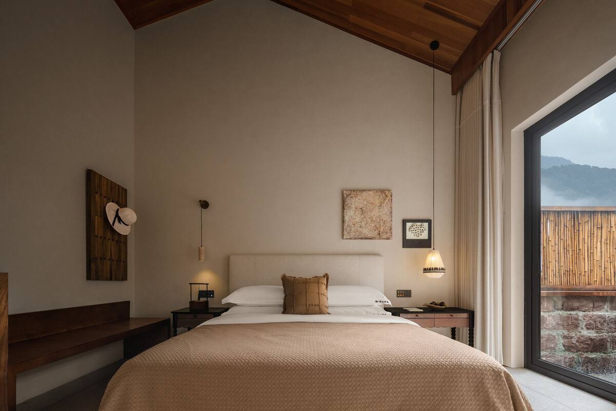 A bedroom with a large bed, wooden headboard, bedside tables, a wall-mounted fan, and a view of mountains through a large window.