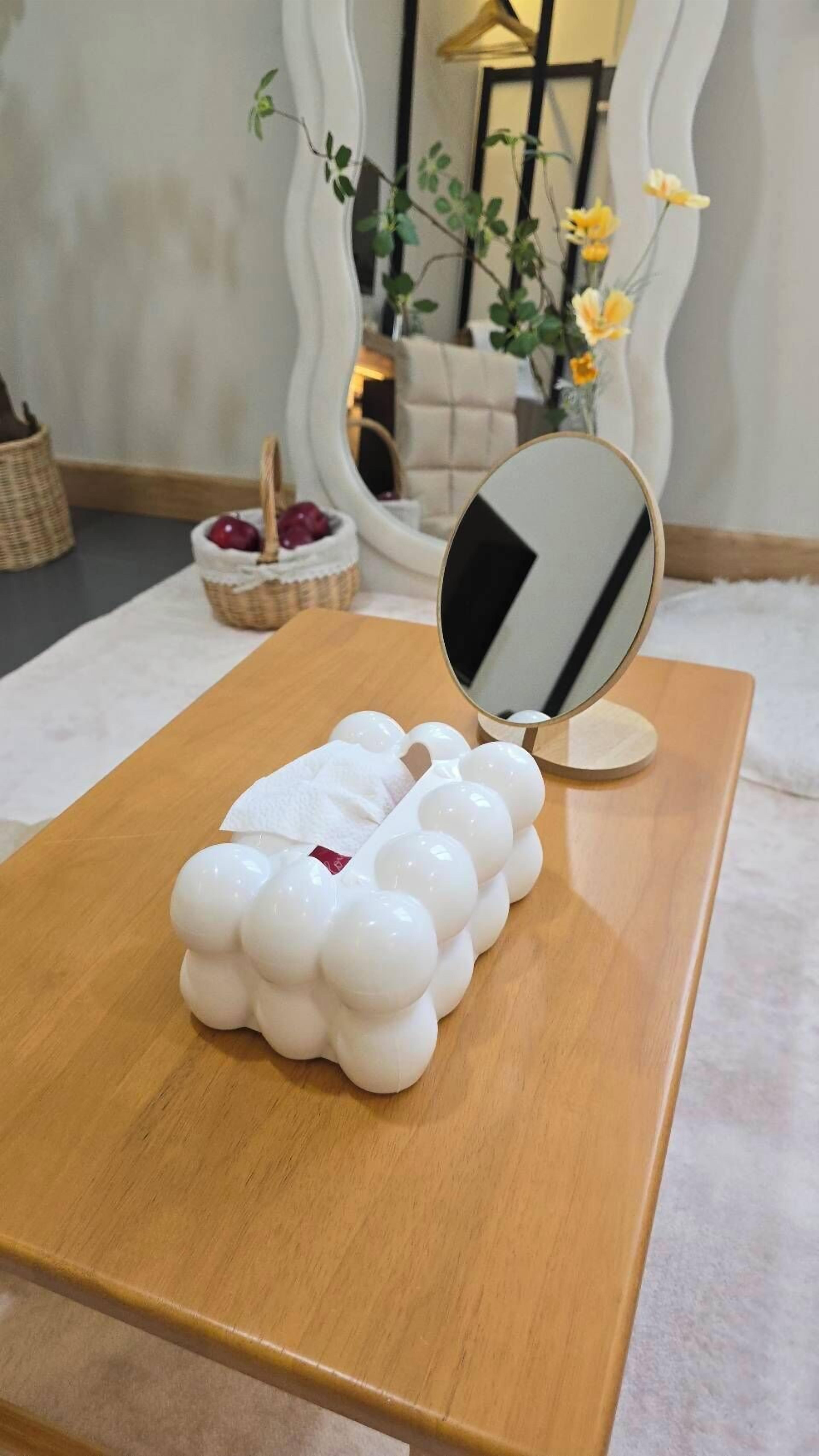 Uma mesa de madeira com um objeto branco que lembra uma nuvem, um espelho e uma tigela com frutas.