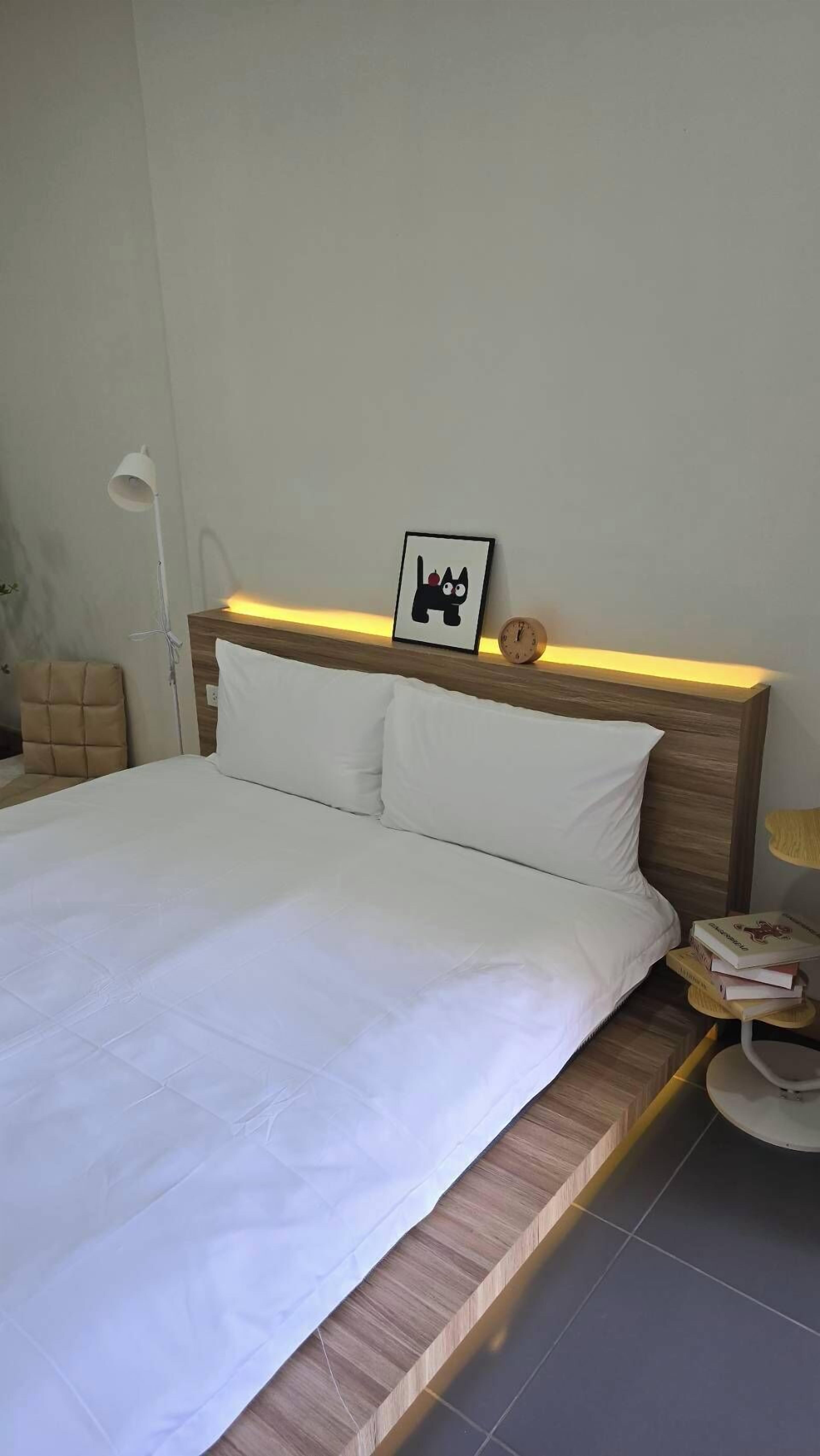 Una cama bien hecha con ropa blanca, un cuadro enmarcado encima, una mesita de noche con un reloj y una lámpara pequeña en la mesita de noche.