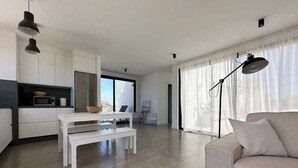 Un saló modern amb un sofà, una taula de dinar amb bancs, una zona de cuina i un finestral gran amb cortines.