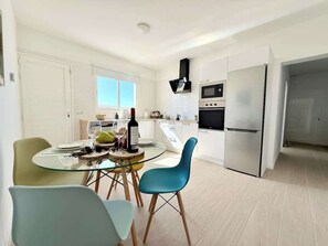 Eine moderne Küche mit weißen Schränken, einem Edelstahlkühlschrank und einem eingebauten Ofen. Es gibt eine Essecke mit einem Glastisch und zwei Stühlen.