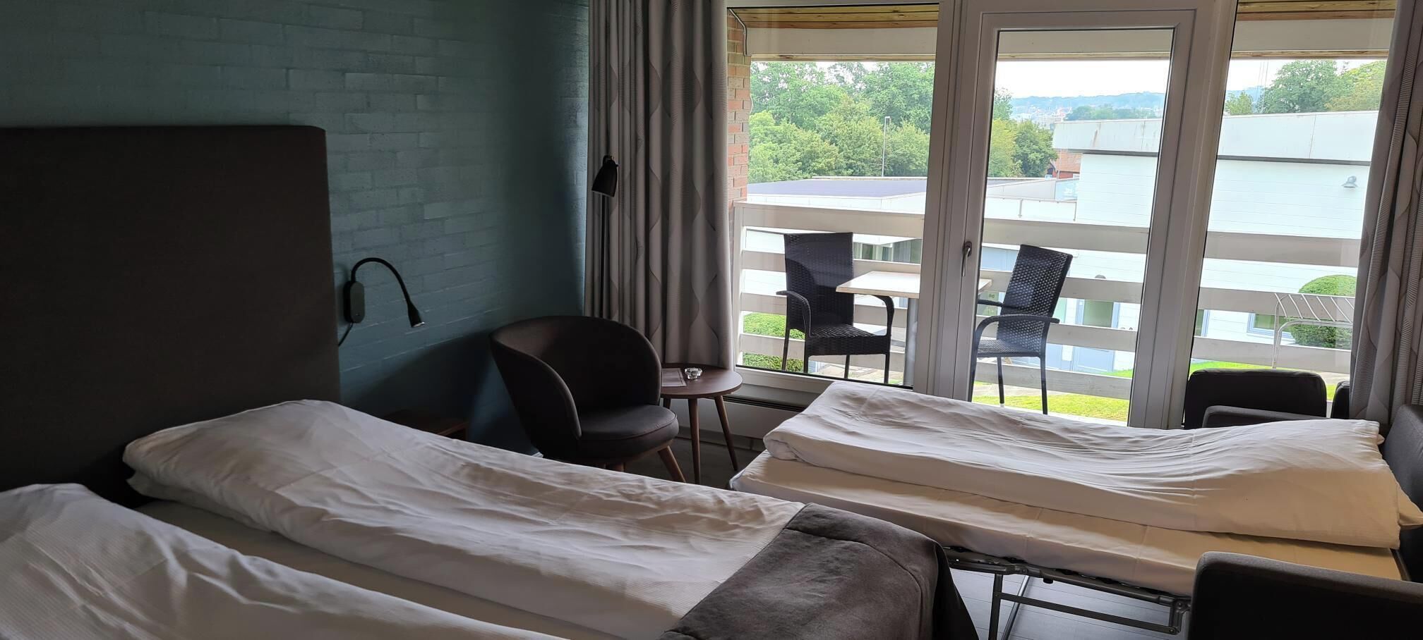 A hotel room with two beds, a small table, a chair, and a balcony with a view of a body of water and greenery.