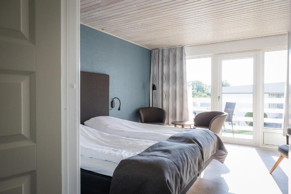 A modern hotel room with a bed, a chair, a small table, and a large window offering a view of the outside.