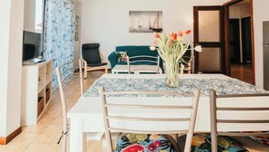 Un comedor con una mesa dispuesta para cuatro, un televisor en un soporte, un sofá y una puerta que da a otra habitación.