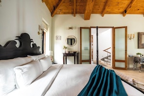 Habitación de hotel con cama, mesita de noche, espejo, una pequeña mesa con lámpara y vista a una escalera que lleva a un balcón.