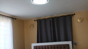 A room with a tufted headboard, a ceiling light, a window with curtains, and a wall-mounted thermostat. - Stacy's Cozy Bed and Breakfast in Nakuru, Kenya (Nakuru)