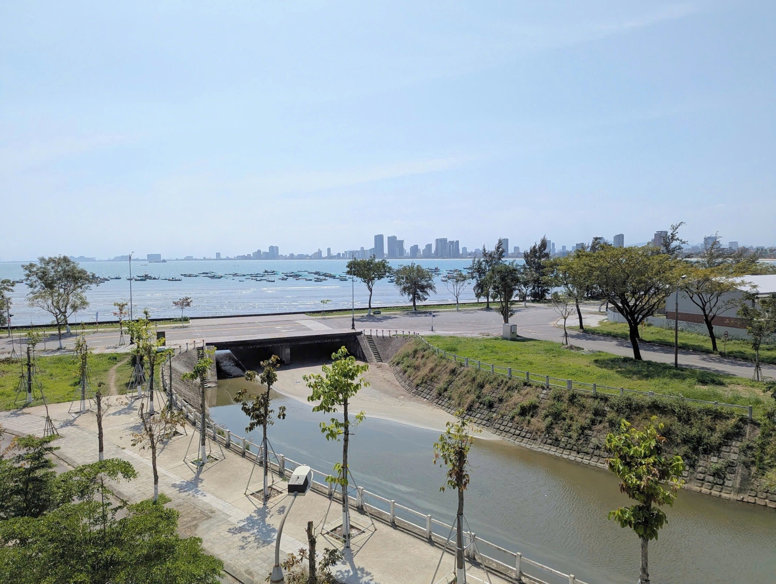 A riverside walkway with trees, a bridge, and a city skyline in the background.