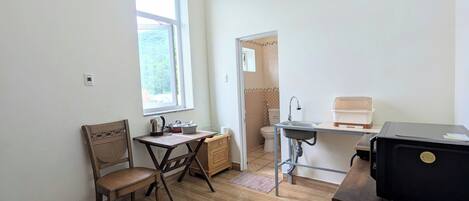 Una habitación pequeña con una silla de madera, una mesita, un ventanal, un lavabo y un electrodoméstico negro.