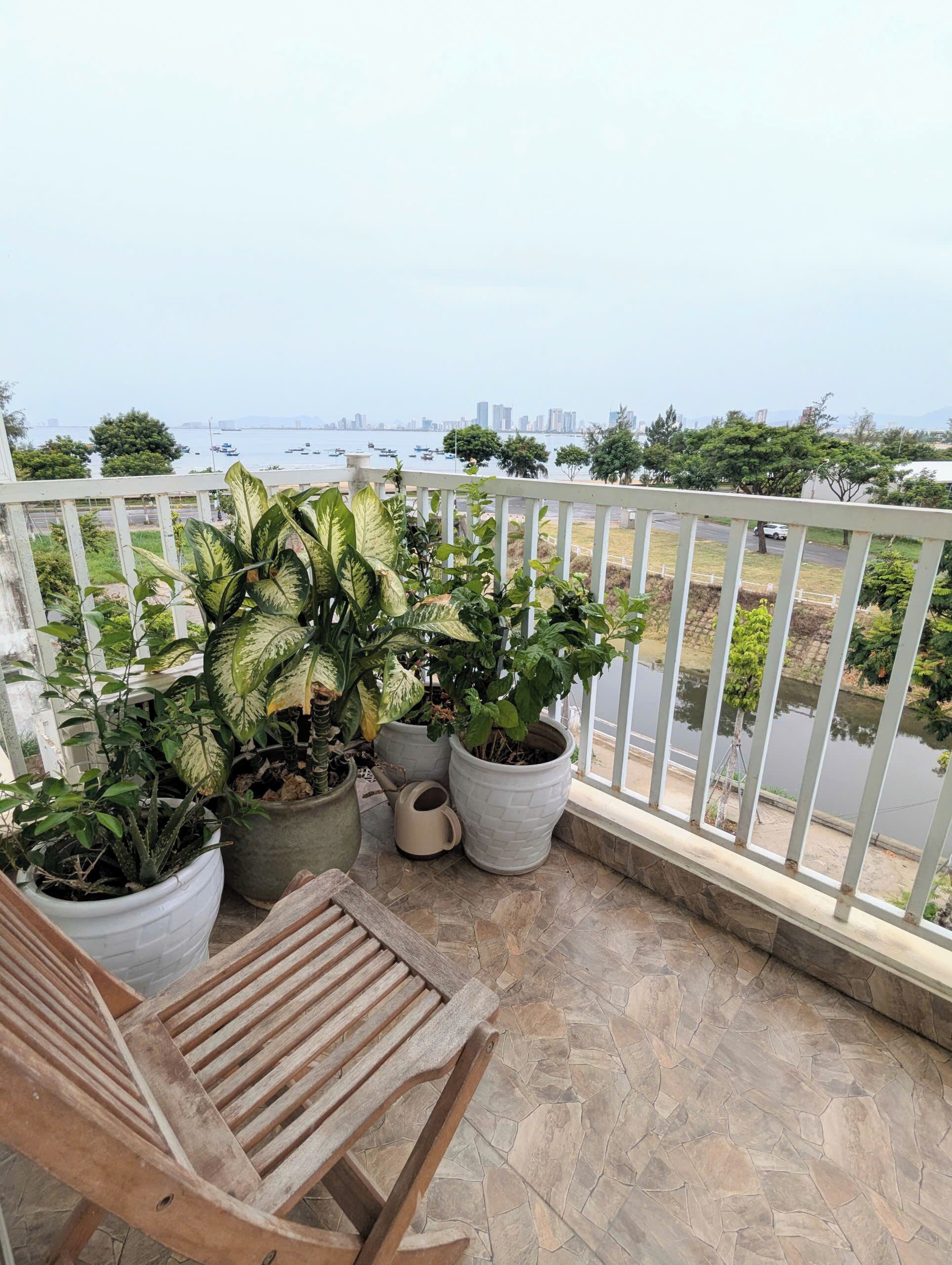 A balcony with potted plants, a wooden chair, and a view of a body of water and cityscape.