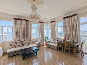 A spacious living room with a sofa, armchair, and dining table, large windows with curtains, and a chandelier. - AN Apartment (Son Tra)
