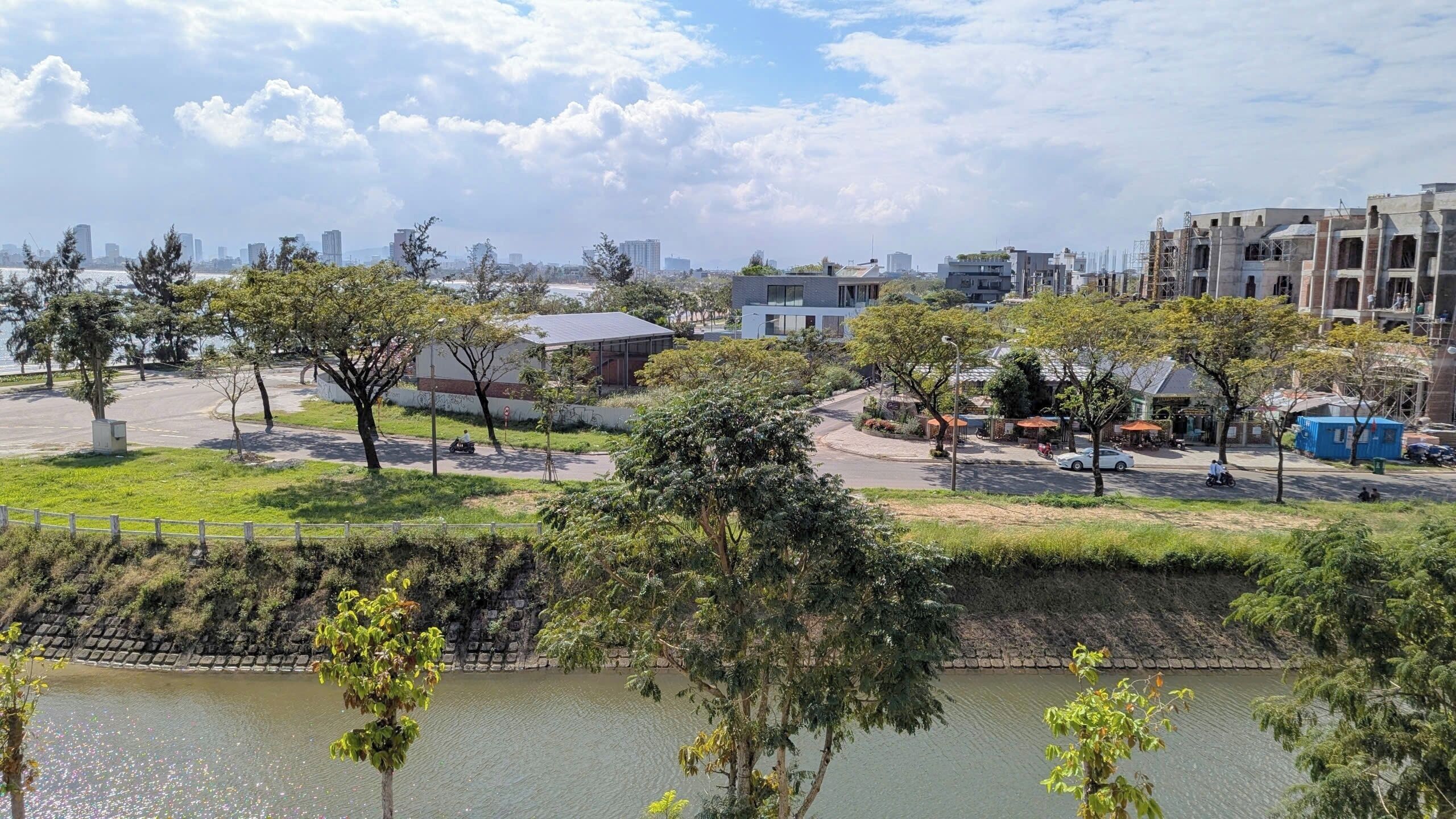 Un parque junto al río con árboles, un sendero para peatones y edificios al fondo.