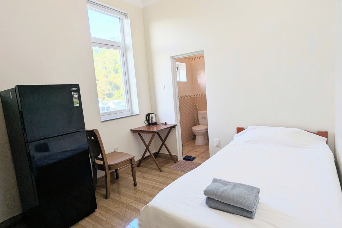 A room with a bed, a desk, a chair, a refrigerator, and a window.
