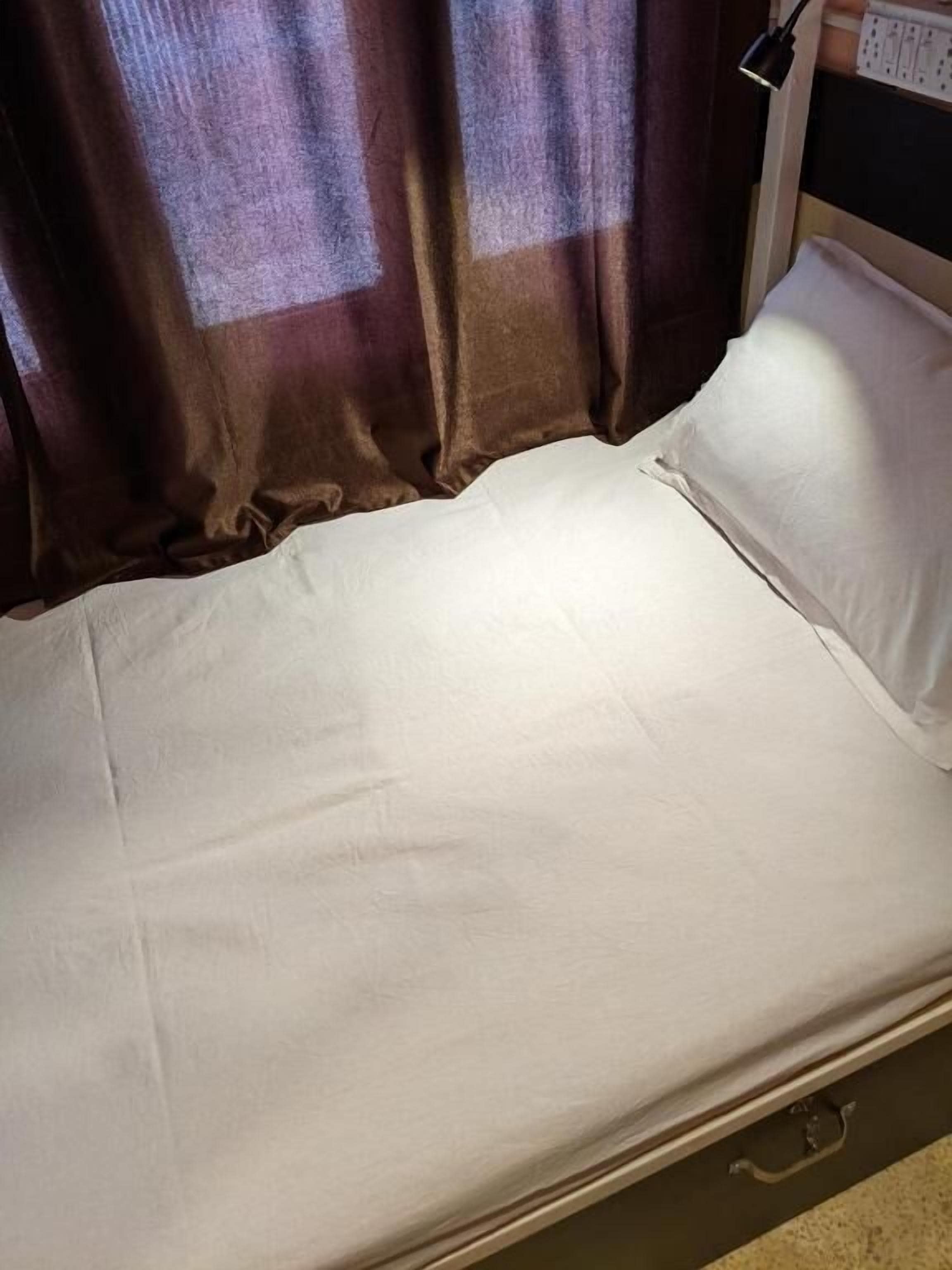 A single bed with a white mattress and pillow, a bedside table with a lamp, and a power outlet on the wall.