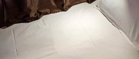 A single bed with a white mattress and pillow, a bedside table with a lamp, and a power outlet on the wall.
