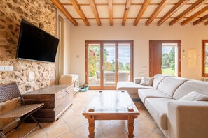 Sala de estar com parede de pedra, vigas de madeira no teto, televisão de tela plana, sofá, mesa de centro e porta de vidro deslizante que dá para uma área externa.