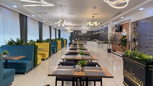 Een modern restaurant met lange tafels, blauw-geel zitmeubilair en decoratieve verlichting.