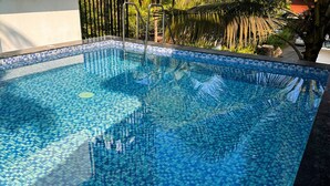 A rectangular pool with a blue mosaic tile pattern, surrounded by palm trees and a white wall. - CELESTE VILLA (Candolim)