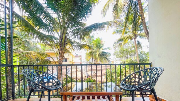 A balcony with a wooden bench and two wicker chairs, overlooking a residential area with palm trees and buildings. - CELESTE VILLA (Candolim)