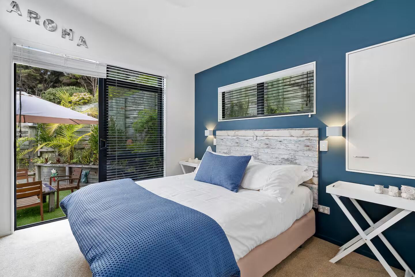 A bedroom with a bed, a nightstand, a window with blinds, and a view of an outdoor patio.