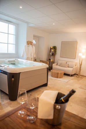 Une salle de spa avec un bain à remous, une table en bois avec des verres à vin et une bouteille, un canapé, une échelle ornée de plantes et une œuvre d’art encadrée accrochée au mur.