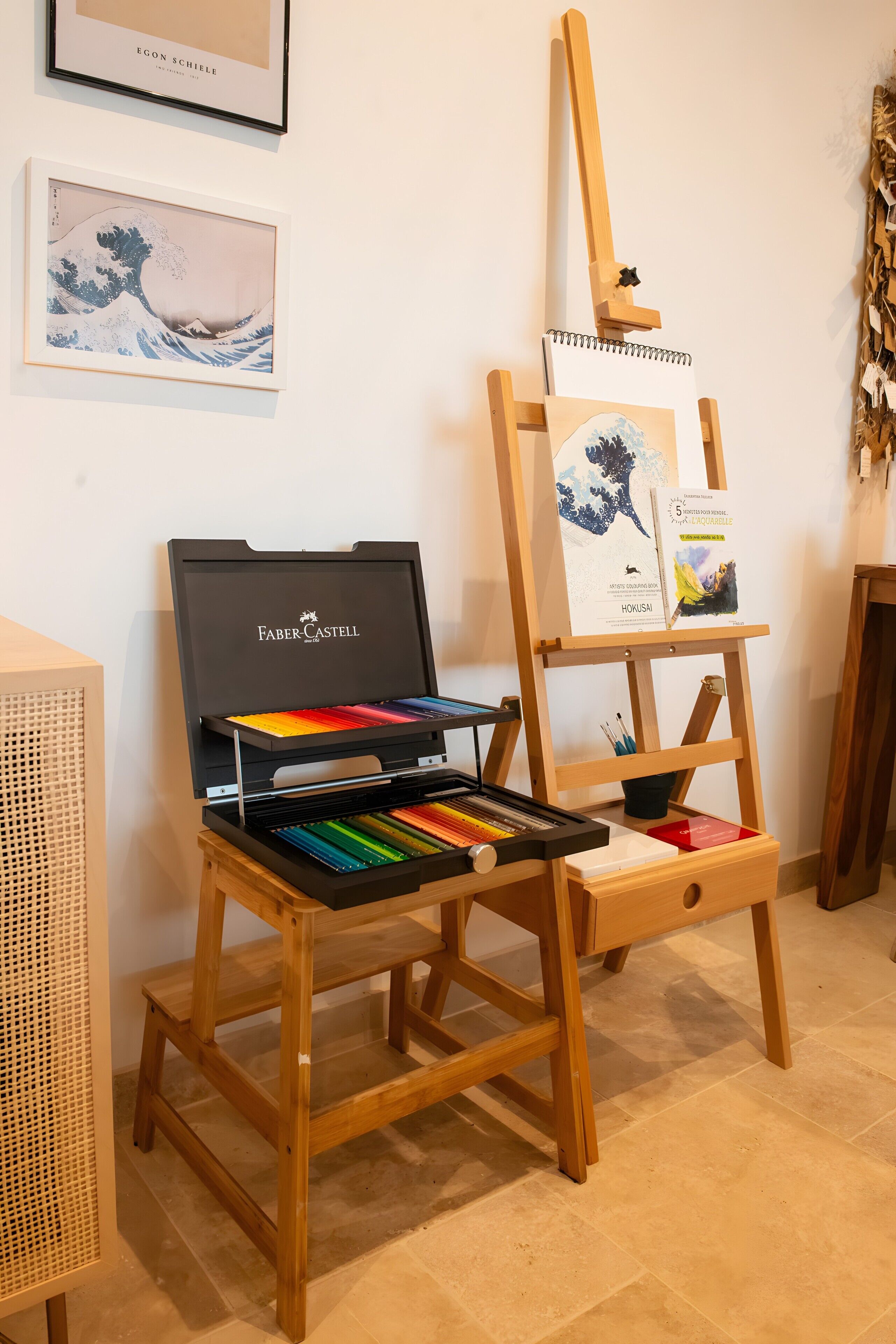 Ein Staffelei mit einem Gemälde, ein Faber-Castell Kunstset und ein Buch auf einem Regal.