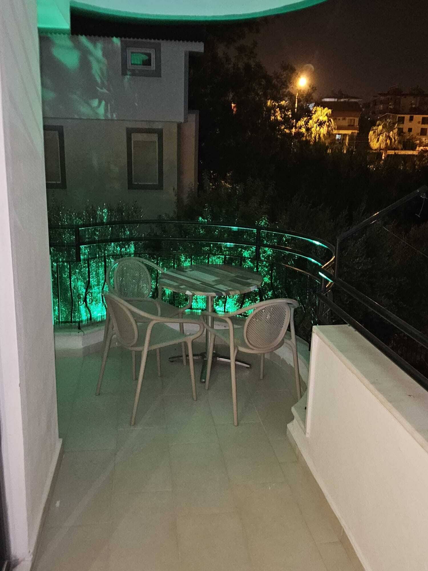 An outdoor balcony with a table and chairs, a railing, and a view of the surrounding buildings at night.
