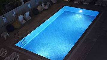 A rectangular swimming pool with a tiled deck, surrounded by lounge chairs and a small deck area with a patterned umbrella.