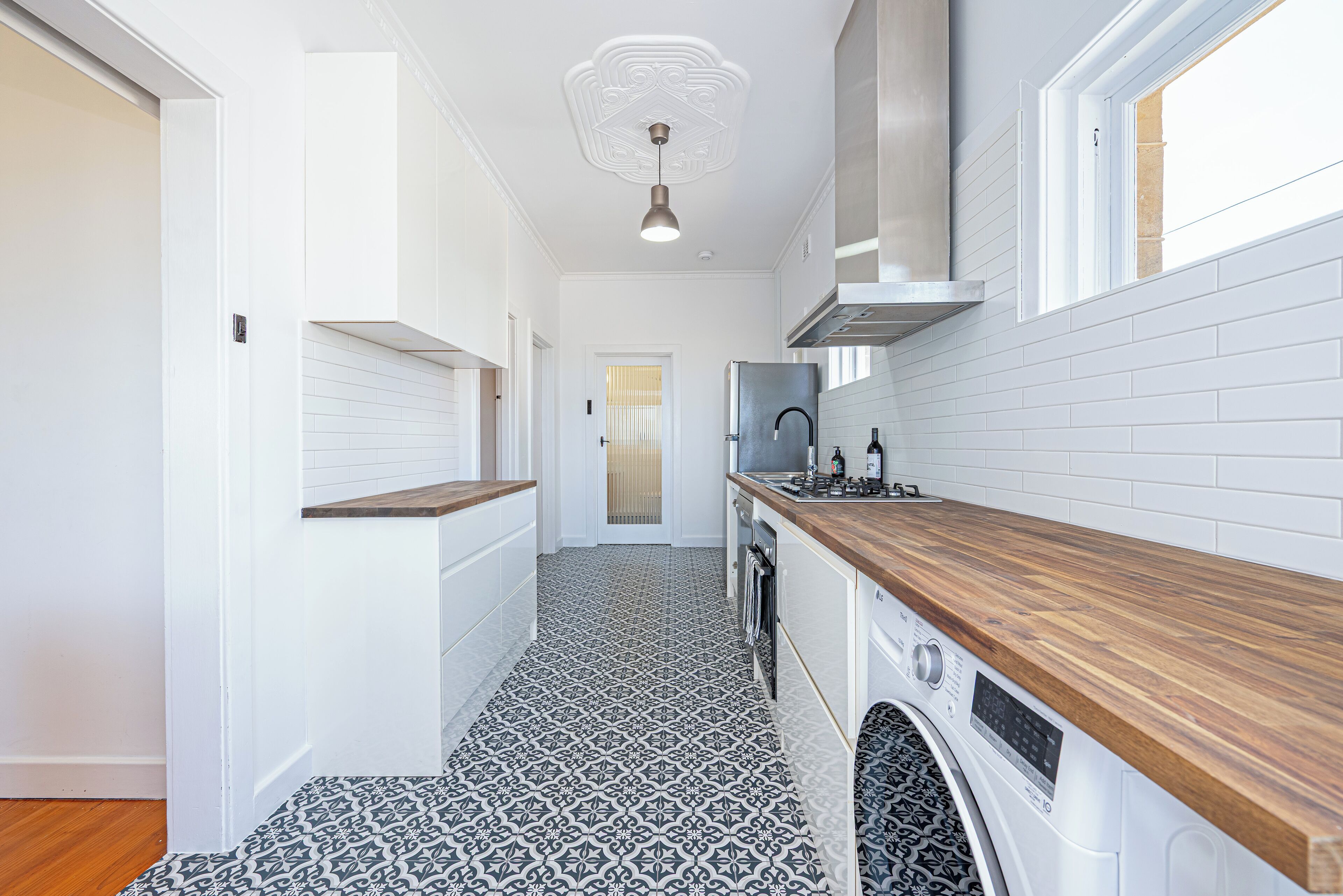 A narrow kitchen with white tiled walls, a patterned floor, and a wooden countertop.