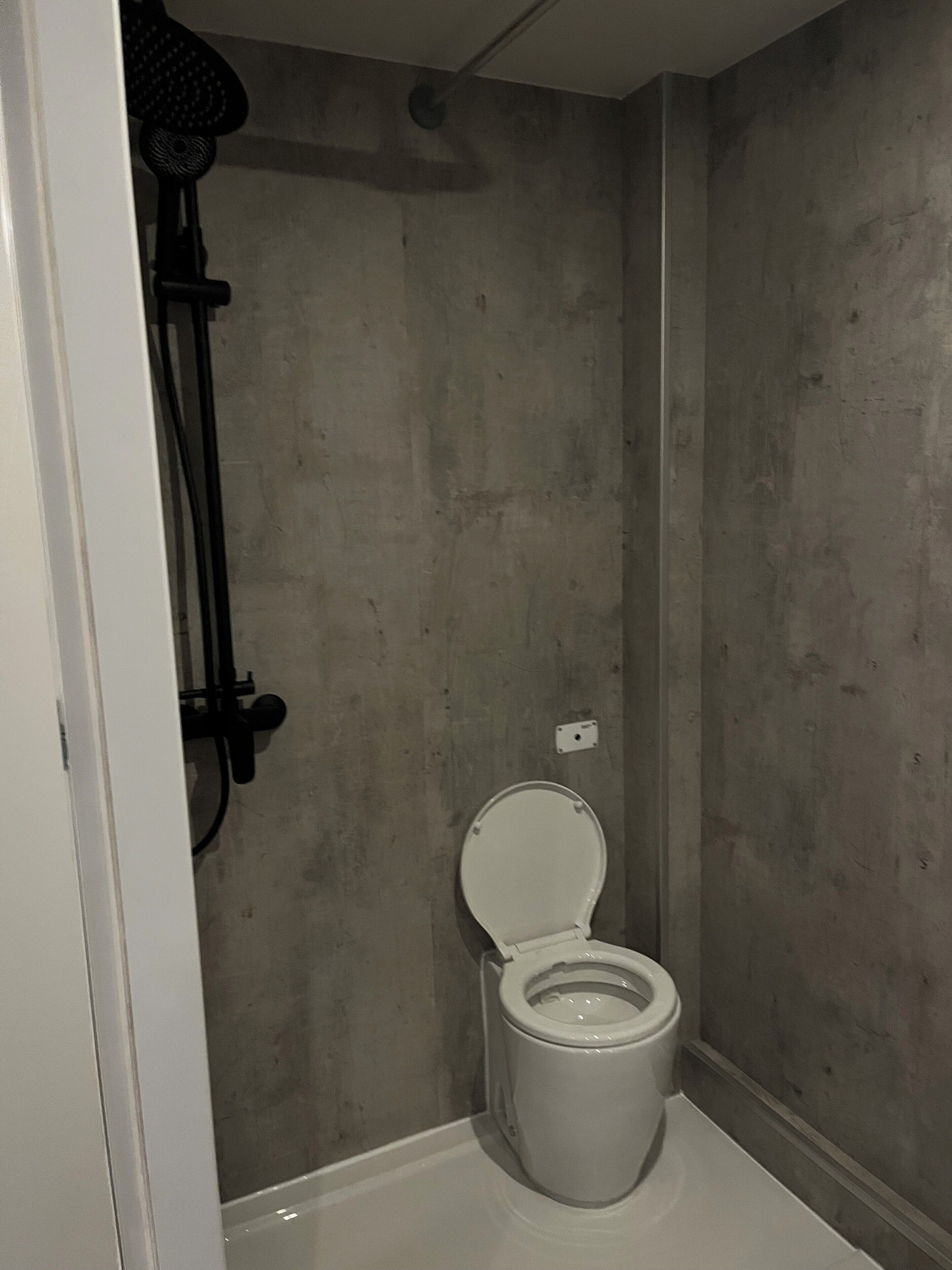 A small, narrow bathroom with a toilet, a shower head, and a wall-mounted light fixture.