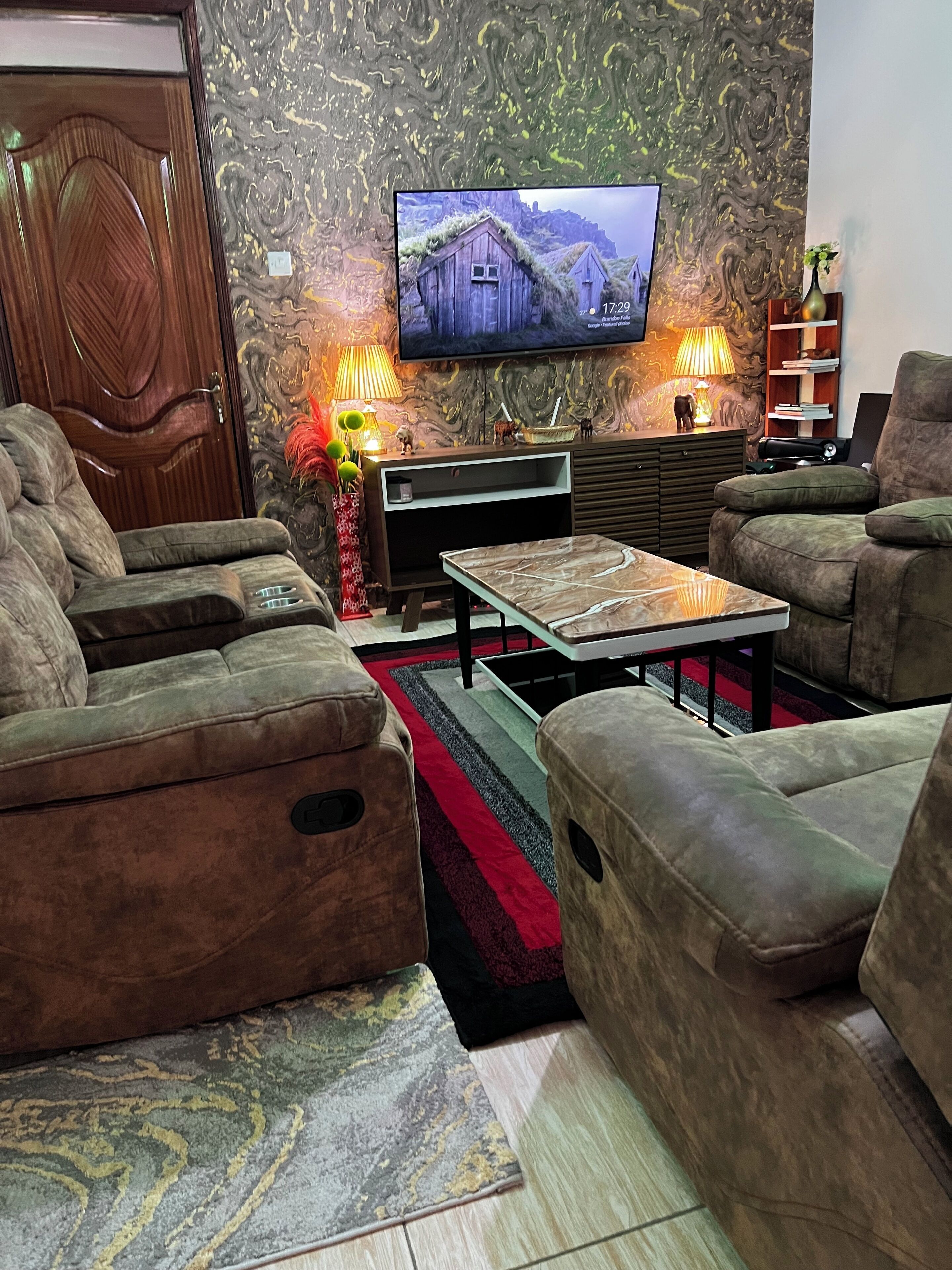 A living room with a large flat-screen TV mounted on the wall, a sofa set, a coffee table, and a patterned rug.