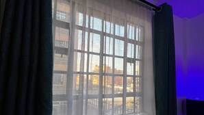 Habitació amb llit, finestra amb cortines lleugeres i vista a edificis.