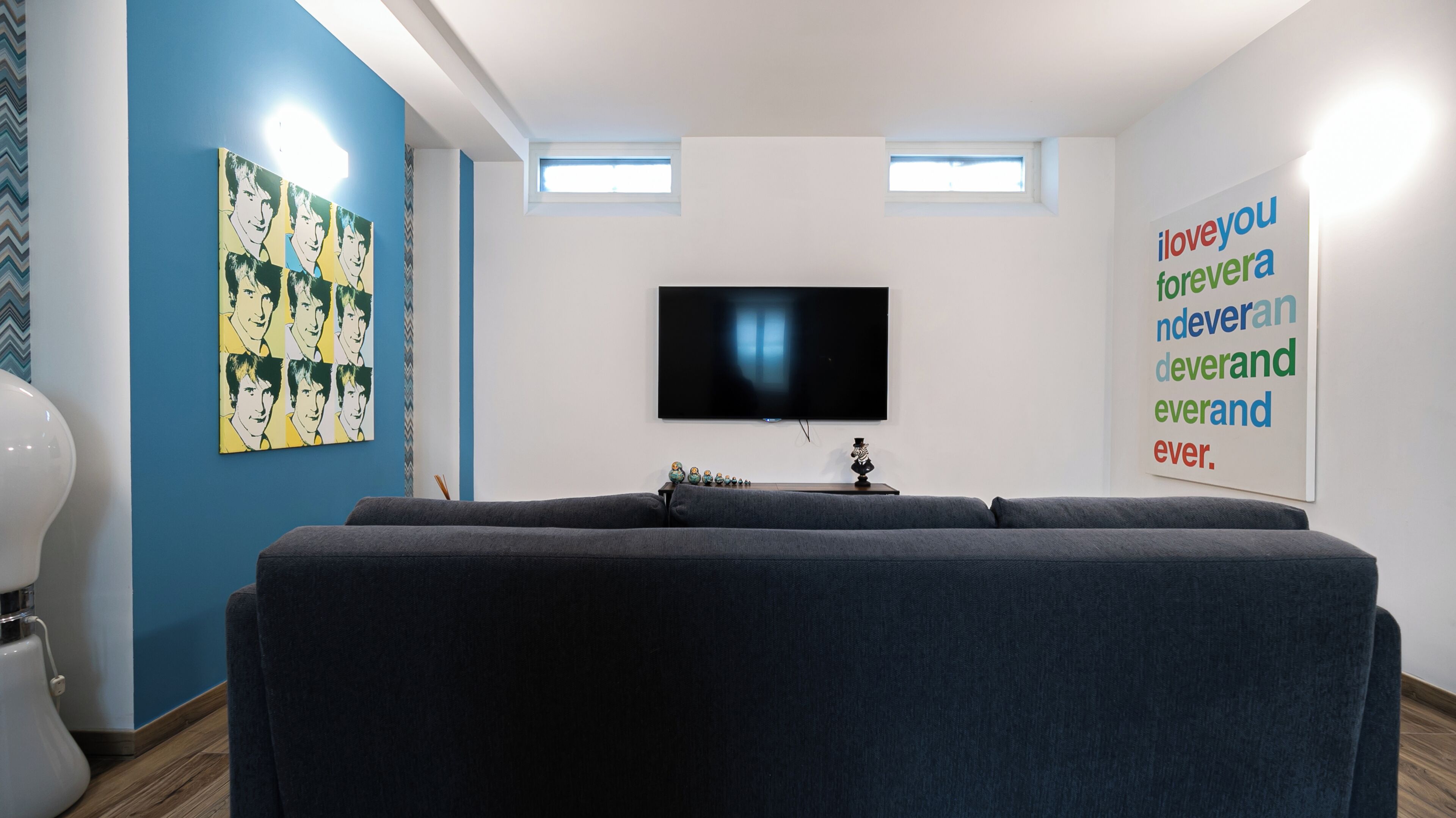 A modern living room with a large grey sofa, a flat-screen TV mounted on the wall, and a colorful poster on the blue wall.