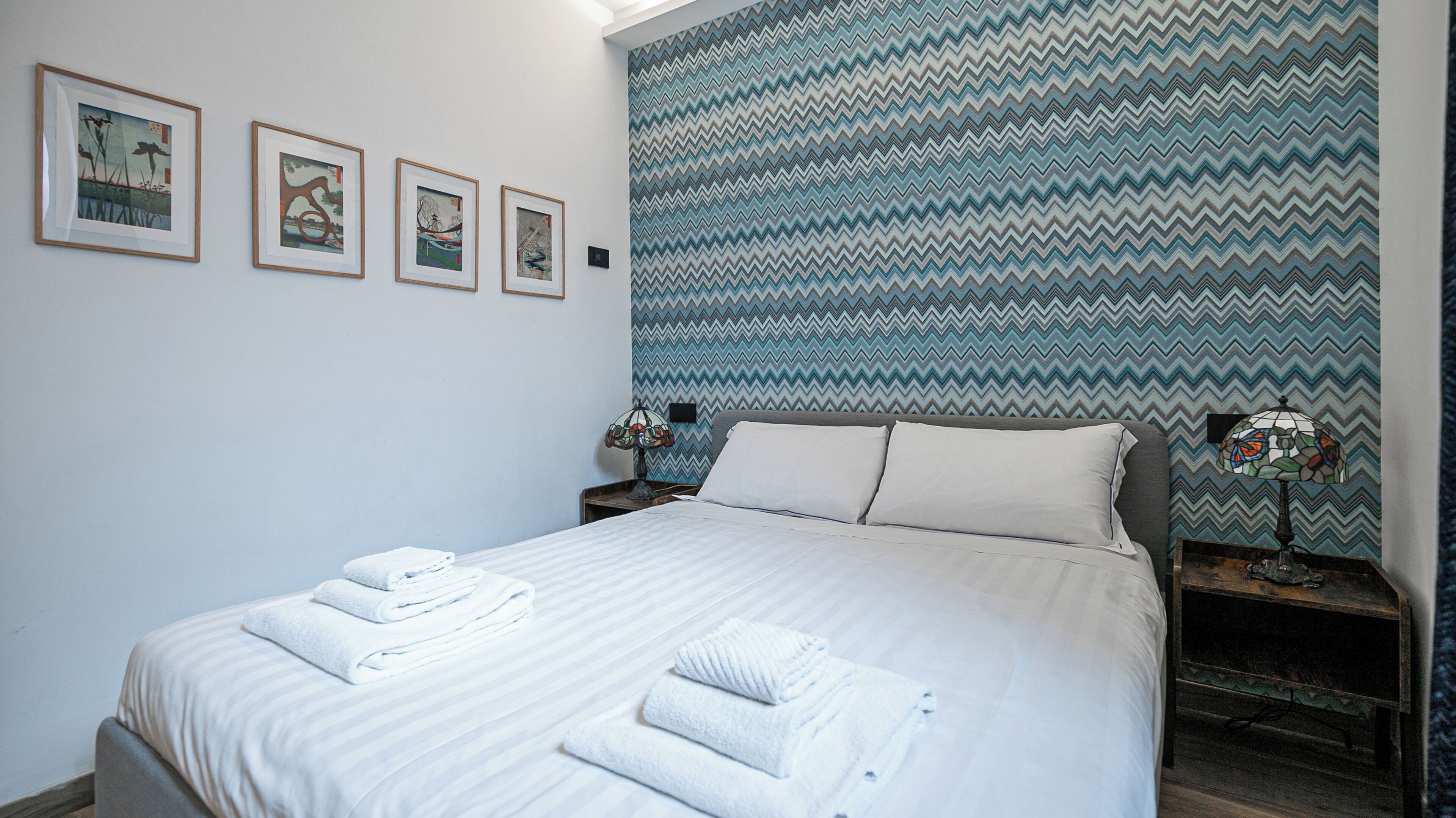 A neatly made bed with folded towels, a patterned wall, and framed artwork on the wall.