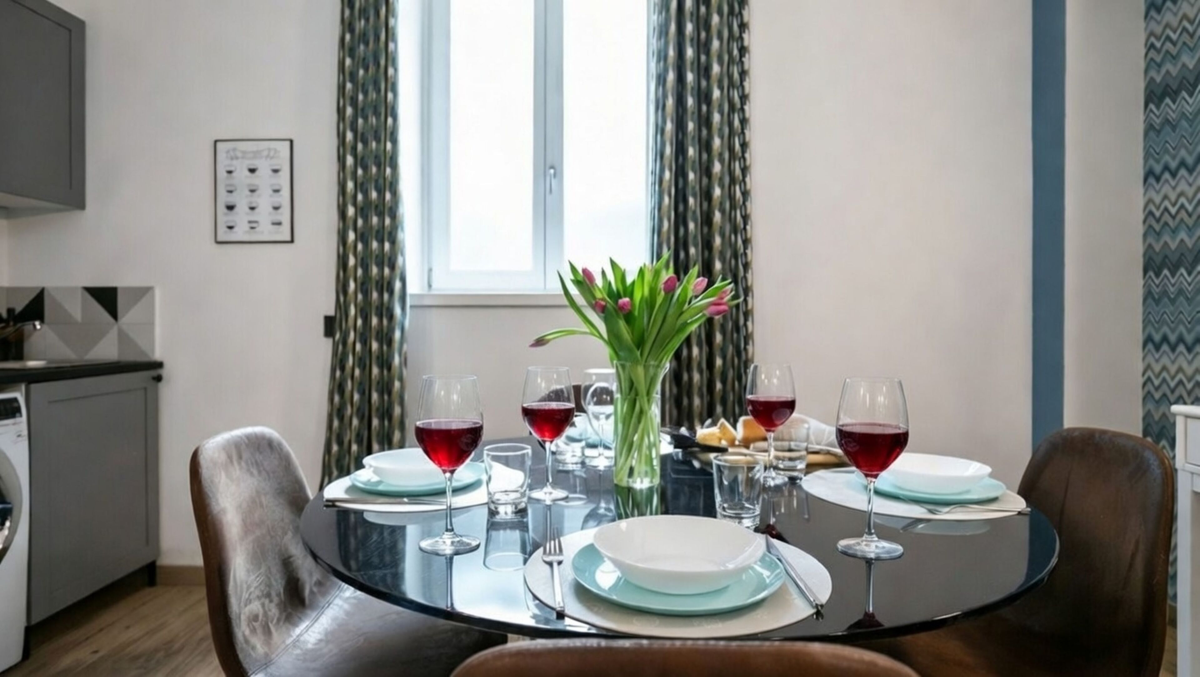 A dining area with a round table set for four, surrounded by chairs. There are wine glasses and plates on the table, and a vase with flowers.