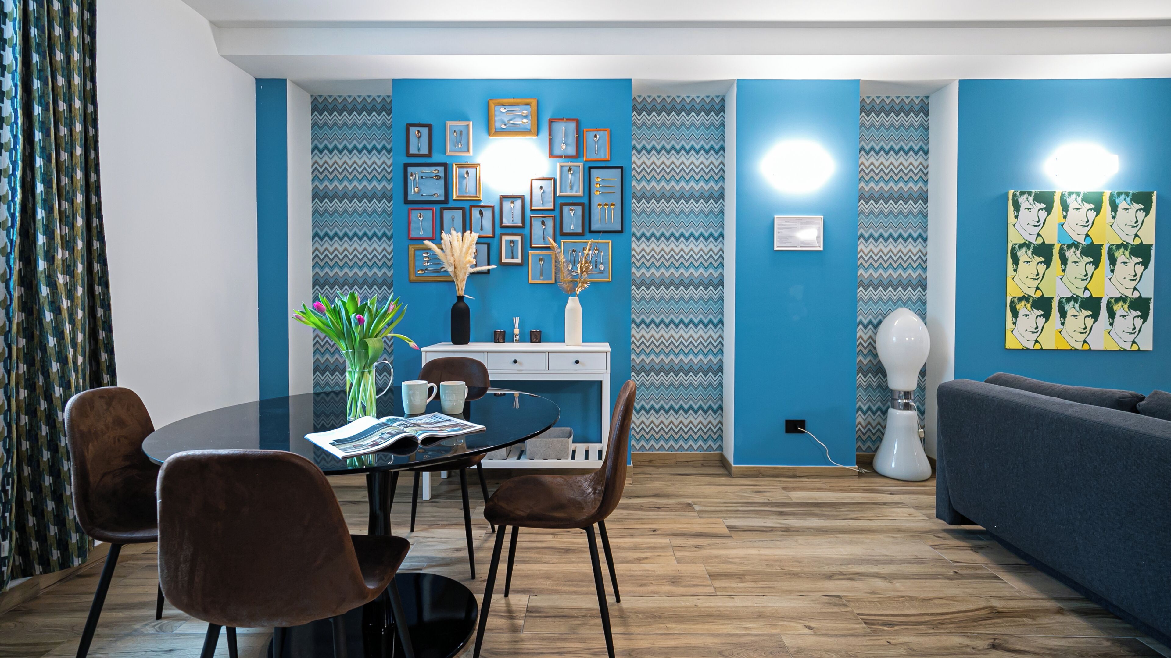 A modern living room with a blue accent wall, a dining area with a round table and chairs, and a grey sofa.