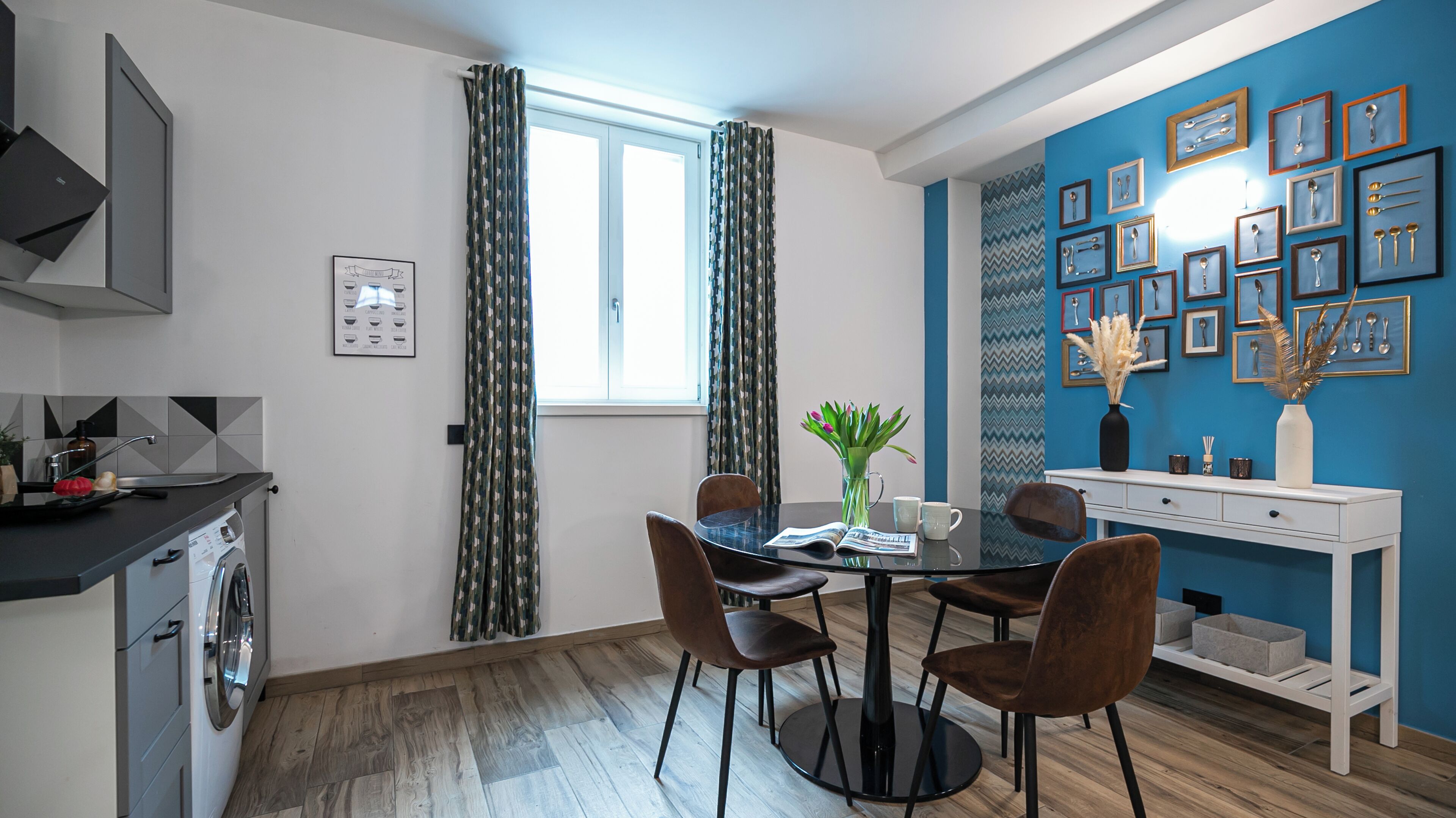Cocina compacta con lavadora, zona de comedor con mesa y sillas, y pared azul decorada con utensilios enmarcados.