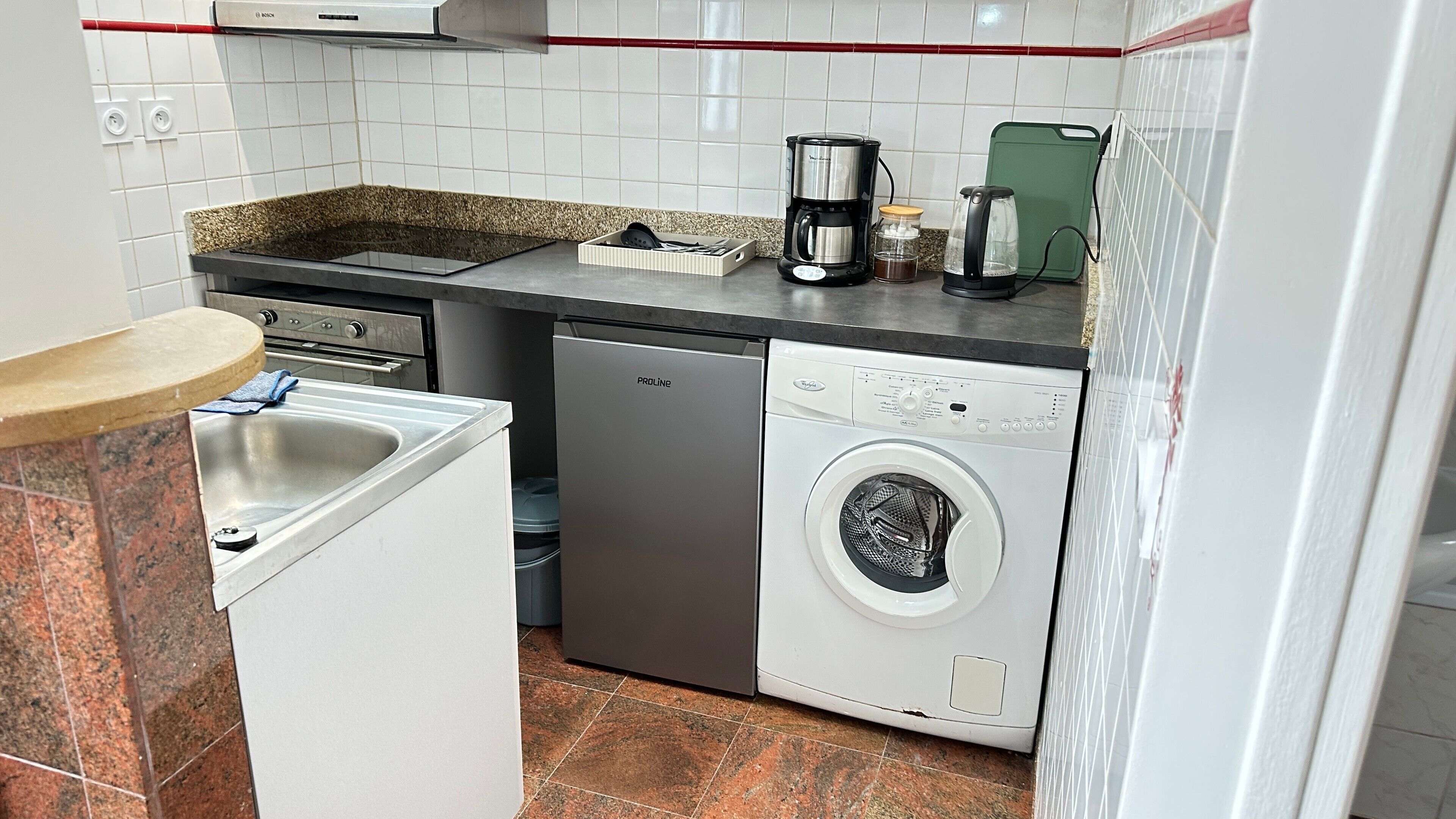 A compact kitchen with a dishwasher, a coffee maker, and a green cutting board.