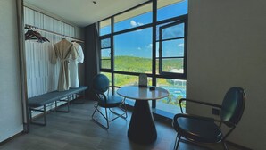 Ein Zimmer mit einem großen Fenster, einem runden Tisch, zwei Stühlen, einer Bank und einem Kleiderständer mit Kleidungsstücken.